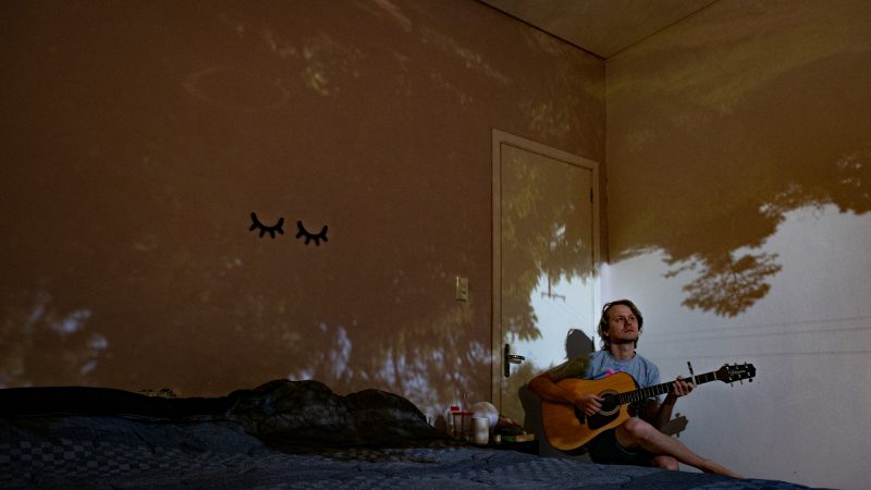 Josué Braun assis seul sur son lit, joue de la guitare. La pièce est sombre et des images d'arbres sont projetées sur les murs.
