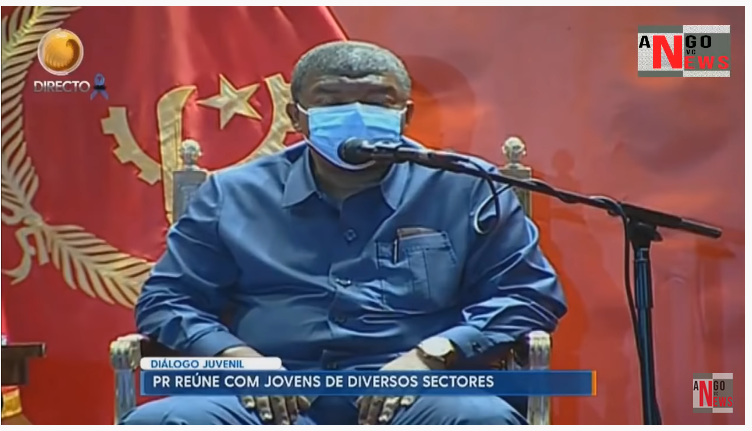 Un homme âgé en costume bleu s'exprime au micro, devant le drapeau de l'Angola. Il porte un masque de protection faciale.